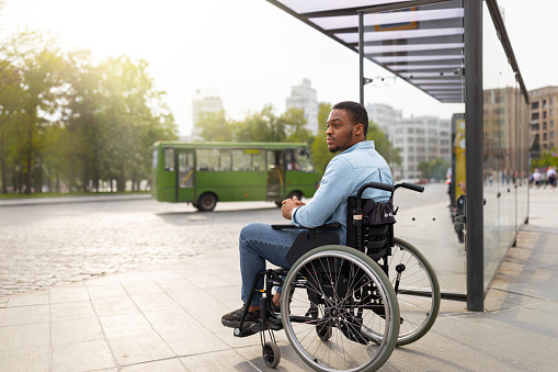 Young disabled black man in wheelchair waiting for public transport on bus stop, having difficulty traveling around city, copy space. Millennial handicapped guy at urban auto station