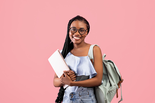 Academic education. Young black woman with backpack and notebooks smiling at camera on pink studio background. Young female university student with afro bunches ready to start school