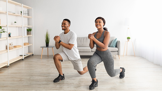 Home fitness. Full length of motivated black couple doing lunges in living room, banner design. Handsome black guy and his girlfriend working out together, training leg muscles during coronavirus