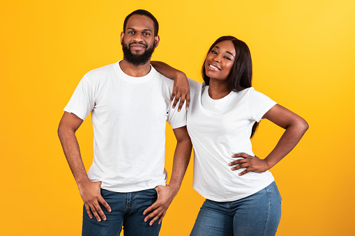 Confident People. Portrait of smiling millennial black woman leaning on her man's shoulder, couple in casual white t-shirts standing and posing isolated over yellow orange studio background