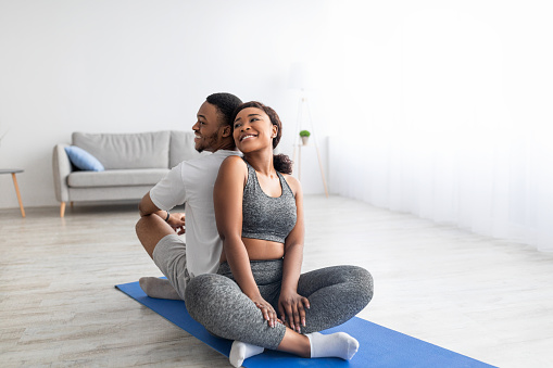 Cheerful black lady sitting back to back with her sporty boyfriend in lotus position, having domestic partner yoga practice. Positive young couple working out together during coronavirus