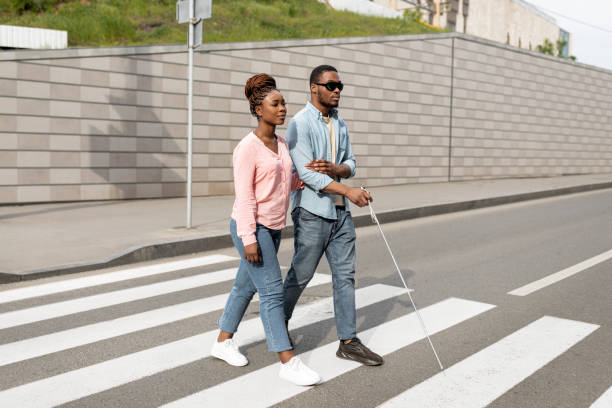 Young black woman assisting visually impaired millennial guy with cane crossing city street. Vision disability concept Young black woman assisting visually impaired millennial guy with cane crossing city street. African American blind man with his guide using crosswalk in downtown area. Vision disability concept blindness stock pictures, royalty-free photos & images