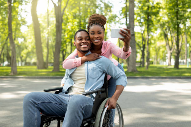 mujer negra feliz y su novio discapacitado en silla de ruedas tomándose selfie juntos, abrazándose en el parque de la ciudad - silla de ruedas fotografías e imágenes de stock