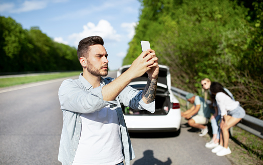 Upset young guy standing on road next to broken car, looking for mobile network signal, trying to call roadside assistance, having no connection, his friends trying to fix flat tire, copy space