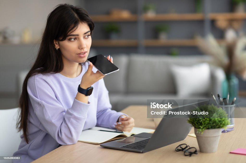 Woman using voice search assistant on smart phone Work From Home. Portrait Of Young Business Lady Using Voice Search Assistant, Talking On Mobile Phone And Using Laptop Computer. Woman Sitting At Desk With Pc At Home Office, Free Copy Space Filming Stock Photo