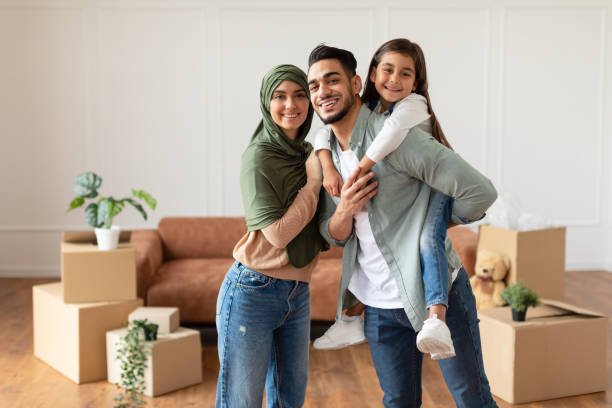 Happy muslim family looking at camera, posing on moving day Portrait of excited muslim man, woman in headscarf and girl posing in new apartment, standing in empty living room with cardboard boxes. Cheerful father carrying his daughter on back modest clothing stock pictures, royalty-free photos & images