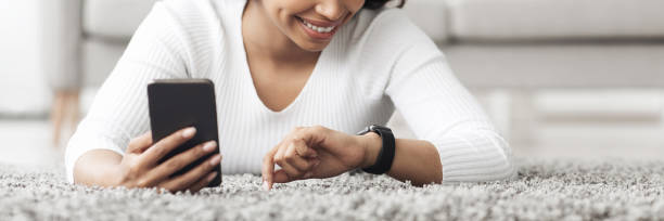 African american woman using smartwatch with e-mail notifier, connecting smartphone to modern hand device, panorama African american woman using smartwatch with e-mail notifier, panorama. Lady connecting smartphone to modern hand device while lying on floor, crop. Technology concept Notifier stock pictures, royalty-free photos & images