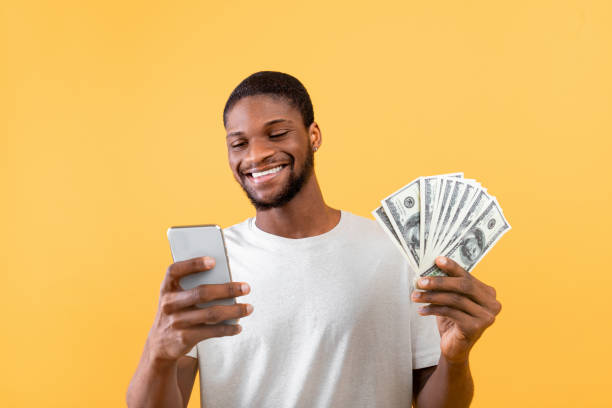 istock Make money online and cashback concept. Happy black guy using smartphone, showing bunch of money, yellow background 1325282048