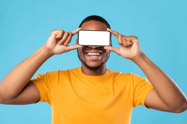 African Guy Holding Smartphone In Front Of Eyes, Blue Background