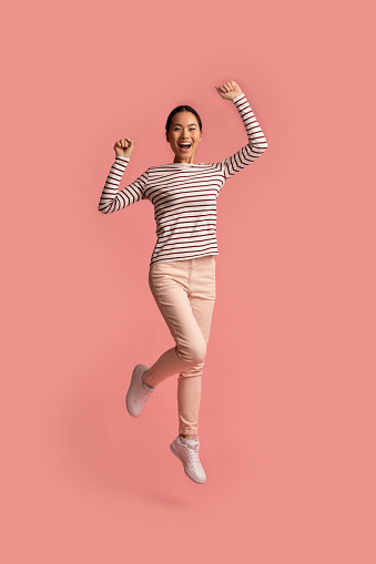 Euphoric Young Asian Woman Jumping With Raised Hands Over Pink Background, Full-Length Shot Of Cheerful Excited Korean Female Celebrating Win With Clenched Fists, Exclaiming With Joy, Copy Space