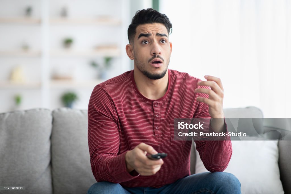 Shocked arab guy wathcing movie on TV Shocked arab guy wathcing movie on TV at home, sitting on couch alone, gesturing, having confused face expression. Speechless young man with TV remote watching horror or thriller, copy space Men Stock Photo