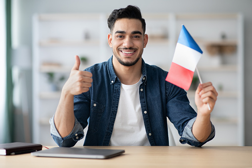 Smiling arab guy with flag of France showing thumb up, sitting at desk with laptop, middle-eastern young man student learning French for job, education or emigration to France, copy space
