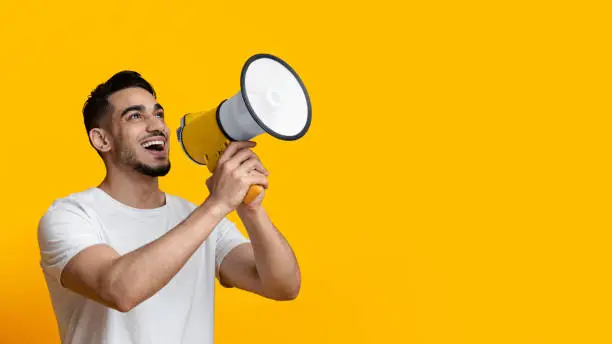 Excited arab guy speaking loud, using megaphone on yellow studio background, panorama with copy space. Happy middle-eastern man give announcement or advertising, using loudspeaker
