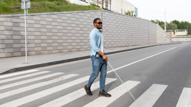 Young black visually impaired man wearing dark glasses, walking across city street, using cane Young black visually impaired man wearing dark glasses, walking across city street, using cane. Millennial blind guy using crosswalk at downtown area, panorama with copy space blind persons cane stock pictures, royalty-free photos & images