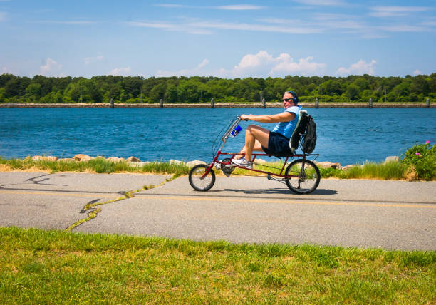 Sandwich, Massachusetts, USA-June 8, 2021- A middle aged man seems relaxed as he wears sunglasses,  shorts and headphones and  peddles his recumbent bicycle along the seven mile paved walkway abutting the Cape Cod Canal.