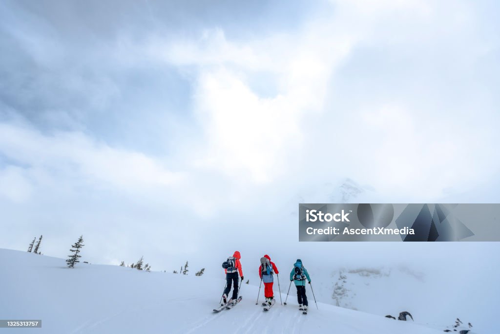 Backcountry skiers traverse snow slope in the morning They climb towards snowy Canadian Rockies, hidden in fog Skiing Stock Photo