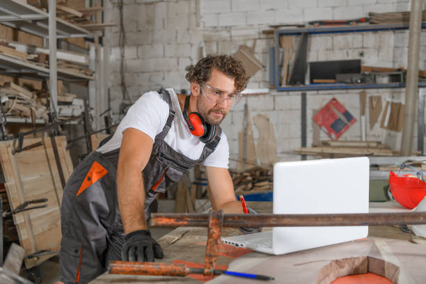 A Male Carpenter in Work Uniform and Protective Workwear is Doing Manufacturing Job in Large Workshop While Using a Laptop for Online Help.