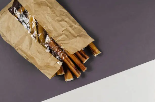 Photo of Pet treats in a paper bag with transparent window. Dental treats on gray and white background. Bully sticks. Beef pizzle for dog. Natural air dried chewing sticks.