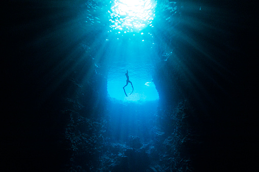 Underwater diver swimming through cave in deep blue ocean towards sunlight