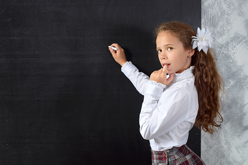 The schoolgirl in school uniform stays at the blackboard, thinks and does the task. Back to school. The new school year. Child education concept.