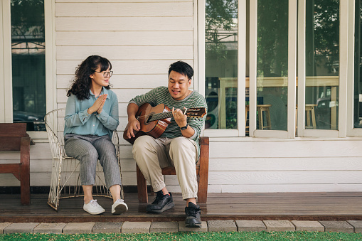 Happy young Asian couple enjoy singing and playing guitar while sitting in front of the house, Couple relaxing and spending time together in backyard at home, People and lifestyle concept