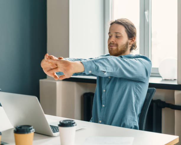 Young tired man office worker in casual clothes stretching arms at workplace