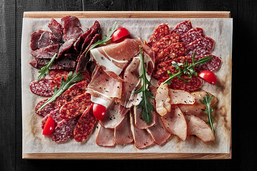 Sliced dry-cured prosciutto, jerked chicken, pork loin and beef fillet, salami and pepperoni on wooden serving board with tomatoes and fresh arugula. Top view of traditional cold cuts. Meat delicacies