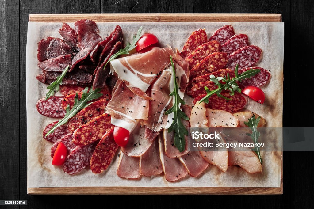 Cold cuts with prosciutto, chicken, pork, beef, sausages Sliced dry-cured prosciutto, jerked chicken, pork loin and beef fillet, salami and pepperoni on wooden serving board with tomatoes and fresh arugula. Top view of traditional cold cuts. Meat delicacies Charcuterie Stock Photo