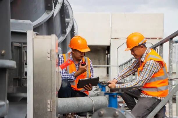 Photo of Asian male engineer inspecting pipe fittings and chillers, refrigeration plant refrigeration plant in  factory. male mechanic wearing helmet and Reflective Safety Vest uses laptop, tablet, equipment with sets of cooling towers in conditioning systems back