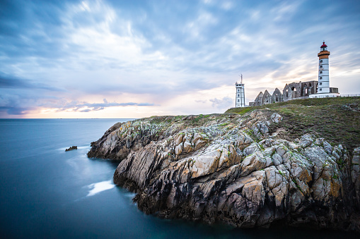 This black-and-white horizontal photograph presents a stunning view of Kannesteinen Beach in Norway, most notably featuring its iconic mushroom-shaped rock. The foreground is covered with sea-eroded stones leading the eye towards the mushroom rock in the middle ground. The calm sea and distant mountain fill the background, all under a dramatically cloudy sky. Captured during a cloudy summer morning, the photograph exudes a sense of stillness and drama.