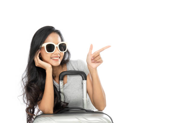 Portrait of an Asian woman wearing a summer dress with a suitcase. She pointed her finger at the copy space on the side. Isolated on a white background. stock photo