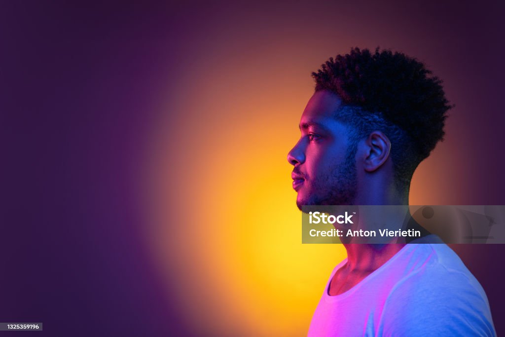 Portrait of a young african man at studio. High Fashion male model in colorful bright neon lights. Art design concept. Portrait of a young pensive african man in profile at studio. Male model isolated in colorful bright neon lights posing on purple background. Side view. People Stock Photo