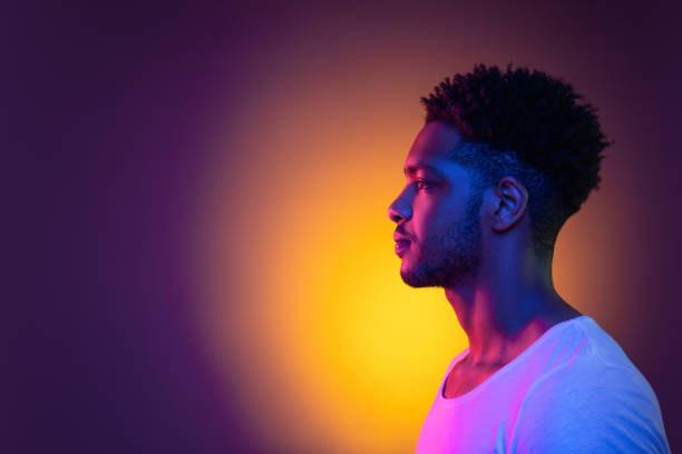 Portrait of a young african man at studio. High Fashion male model in colorful bright neon lights. Art design concept. Portrait of a young pensive african man in profile at studio. Male model isolated in colorful bright neon lights posing on purple background. Side view. artists model photos stock pictures, royalty-free photos & images