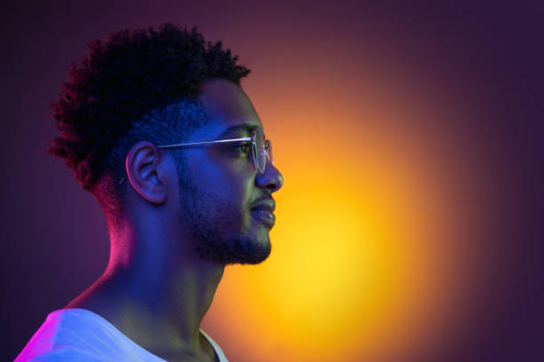 Portrait of a young african man at studio. High Fashion male model in colorful bright neon lights. Art design concept. Close-up portrait in profile of a young african man in glasses posing in colorful bright neon lights on purple background at studio. Side view profile stock pictures, royalty-free photos & images