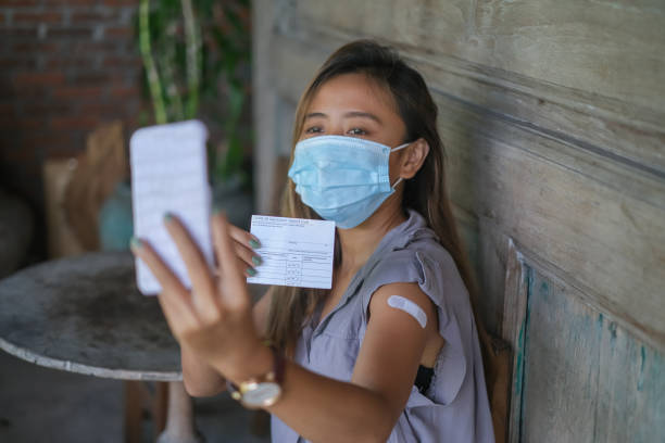 Indonesian Woman Taking a Selfie Showing Her COVID-19 vaccination record card Close-up shot of a proud Indonesian woman with face mask taking a selfie with her smartphone while holding her Covid-19 vaccination record card. immunization certificate photos stock pictures, royalty-free photos & images