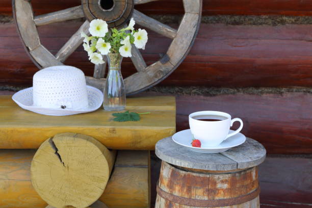 A white mug of black tea on wooden terrace. A summer still life with a mug of black tea, bouquet of flowers in a vase, white hat, wagon wheel, wooden barrel and a yellow bench against a log wall. wagon wheel bench stock pictures, royalty-free photos & images