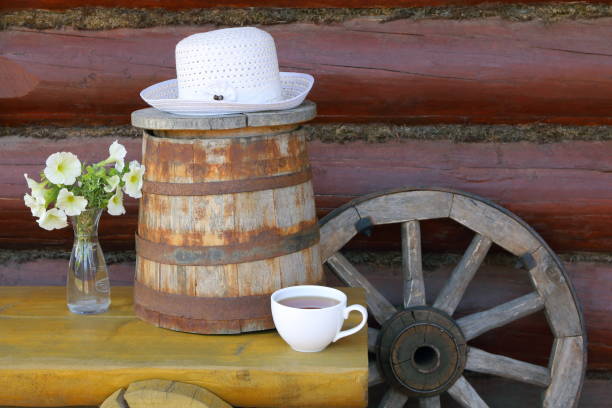 A white mug of black tea on wooden terrace. A summer still life with a mug of black tea, bouquet of flowers in a vase, white hat, wagon wheel, wooden barrel and a yellow bench against a log wall. wagon wheel bench stock pictures, royalty-free photos & images