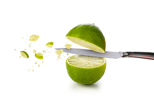 Front view of a lime being cut in halves by a kitchen knife with some lime juice drops and some bites on the air.