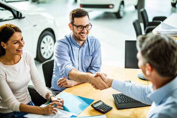Successful deal in a car showroom! Happy couple buying a car in a showroom while men are shaking hands after reaching a successful deal. Focus is on man with glasses. car dealership stock pictures, royalty-free photos & images