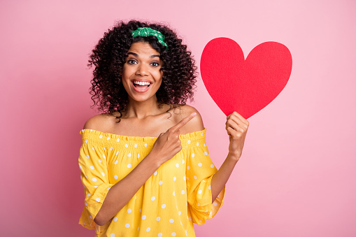 Photo of young african beautiful smiling excited charming girl woman female point finger at big red heart isolated on pink color background.