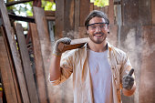 Portrait of woodworker or craftsman wearing safety goggles showing thumb up. Caucasian carpenter holding the wooden bar standing in front of plank background