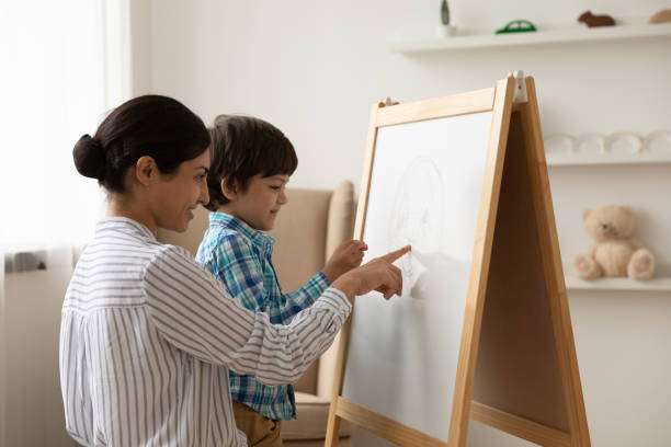 Happy Indian mom and small son draw on whiteboard Loving young Indian mom and little ethnic son draw paint together on flip chart at home. Happy mixed race mother play learn study with small biracial boy child on whiteboard. Funny education concept. kids using smart board stock pictures, royalty-free photos & images