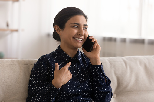 Smiling millennial Indian woman sit relax on sofa at home have pleasant cellphone call with friend or colleague. Happy young mixed race female talk speak on smartphone. Communication concept.