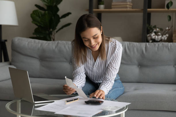 Happy young woman calculating expenditures at home. Happy young woman holding paper bills in hands, calculating expenditures, paying bills or services online in computer application, feeling satisfied with enough money, investment payments concept. energy bill photos stock pictures, royalty-free photos & images