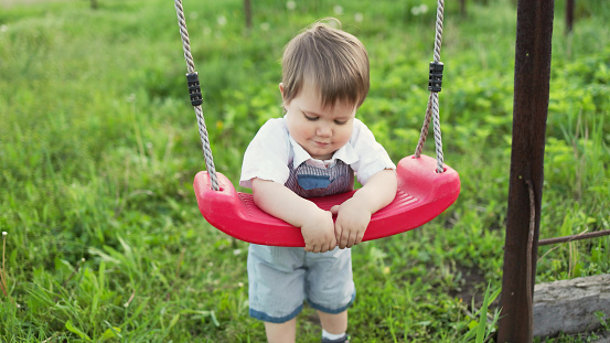 Cute happy baby in a blue summer jumpsuit with bright expressive eyes. Stands near a red street swing in a spring blooming garden covered with greenery.