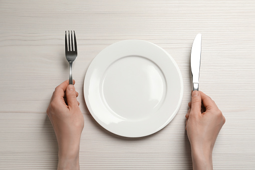 Top view of woman with cutlery and empty plate at white wooden table, closeup