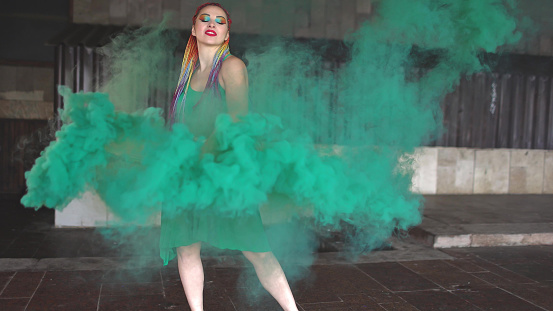A young cheerful girl in a bright green spring dress with makeup with sequins and rainbow African braids. She stands near a building and blows green smoke in the street.