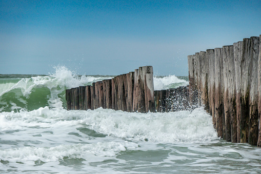 A row of weathered poles stands in the sea to break the waves. The poles are weathered. A large wave washes over the poles. The photograph was taken on the beach at Burgh Haamstede in the Netherlands.