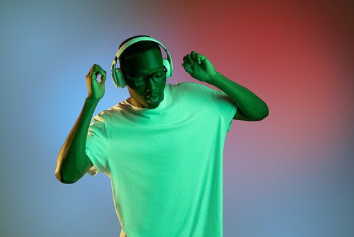 Listening to music. Portrait of young african dancing man in glasses and headphones on bright studio background in neon light. Concept of human emotions, facial expression, youth.
