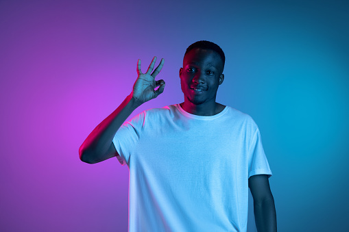 Ok. Smile. Portrait of young smiling african handsome man demostrating ok gesture posing in neon light on bright gradient pink blue background.